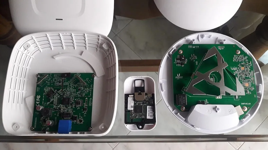 Three access points opened up and laid out on a glass table. Each white plastic housing sits behind its circuit board. On the board of the largest, a flat metal antenna element is visible, shaped and mounted directly on the board. The smallest unit’s board is barely larger than a matchbox.