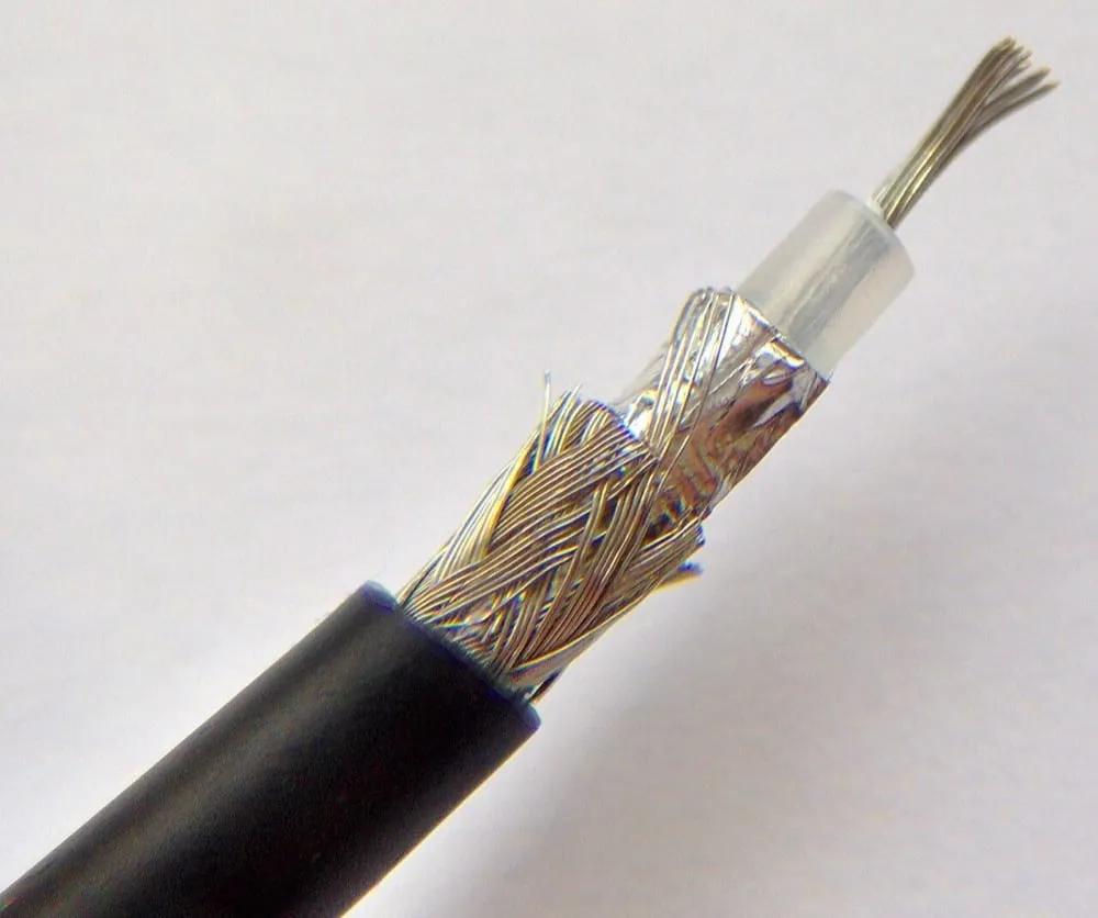 A coaxial cable stripped back in stages so all four layers are visible at once. From the outside in: a black rubber jacket, a woven mesh of fine silver wires, a smooth silver foil beneath the mesh, a clear plastic insulator, and a bundle of bare copper strands at the centre.