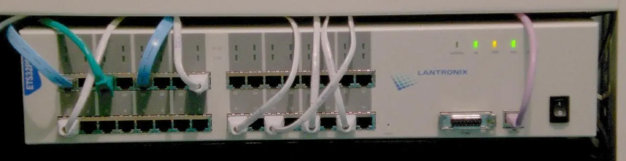 The front of a rack mounted console server photographed straight on. Two rows of RJ45 sockets fill most of the width, thirty two in total, with about a third of them patched with white, blue and green leads that trail out of frame. The manufacturer’s name is printed to the right of the ports, beside a row of status lights, and at the far right are a nine pin serial connector, a single network socket and a power switch.