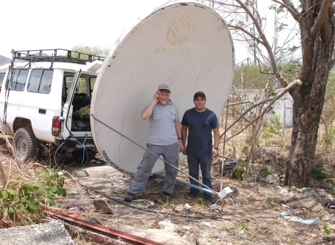 Two people standing in front of a repurposed satellite dish roughly two metres across, mounted at ground level and aimed at the horizon, with a small radio and its feed arm on a pole beside them. Cables run back to a pickup truck.