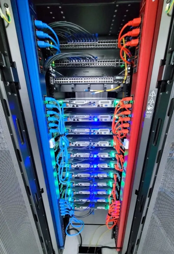 The rear of a full rack, looking down between two vertical power strips. The strip on the left is blue and the one on the right is red. Every server in the rack has two power leads, one blue lead reaching left and one red lead reaching right, so each machine is fed from both strips. Switches and a patch panel fill the top of the rack.