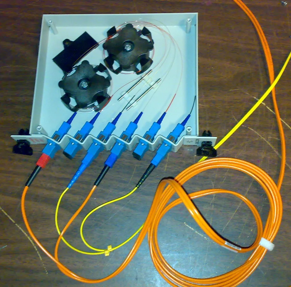 An opened white fibre optic splitter tray sitting on a wooden bench. Two black spools of coiled fibre are mounted inside the tray, with thin fibres routed around them and into the back of six blue SC connectors mounted in a row along the front edge. Orange and yellow patch leads plug into three of the six ports and coil away across the bench. The tray has no power connector.