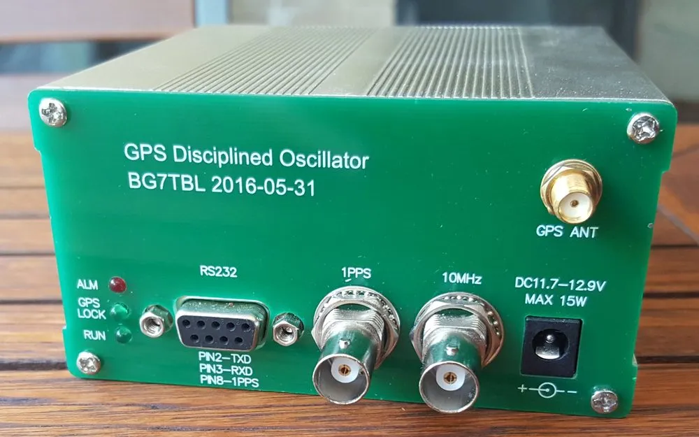 A small green metal box photographed on a wooden table, its front panel silkscreened with white lettering. The panel reads GPS Disciplined Oscillator and carries, from left to right, three indicator lights labelled ALM, GPS LOCK and RUN, a nine pin serial connector labelled RS232 with its pin functions printed underneath, two BNC sockets labelled 1PPS and 10MHz, a power input marked DC 11.7 to 12.9V MAX 15W, and a threaded antenna connector labelled GPS ANT.