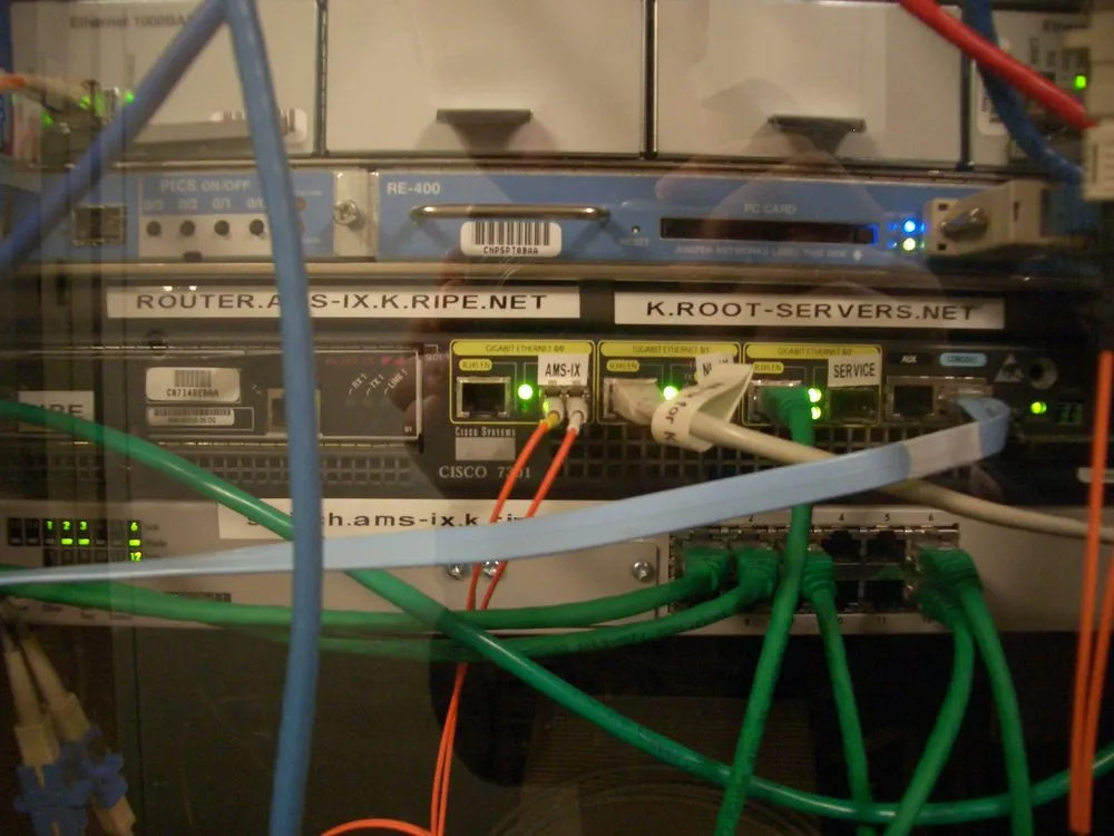 A rack of network equipment photographed through a glass door, with reflections across the image. Two printed labels are fixed to the front panels: ROUTER.AMS-IX.K.RIPE.NET on the left and K.ROOT-SERVERS.NET on the right. Below them a Cisco 7301 router shows three gigabit Ethernet ports with green link lights, and blue, green and orange patch leads run untidily across the front of the equipment in every direction.