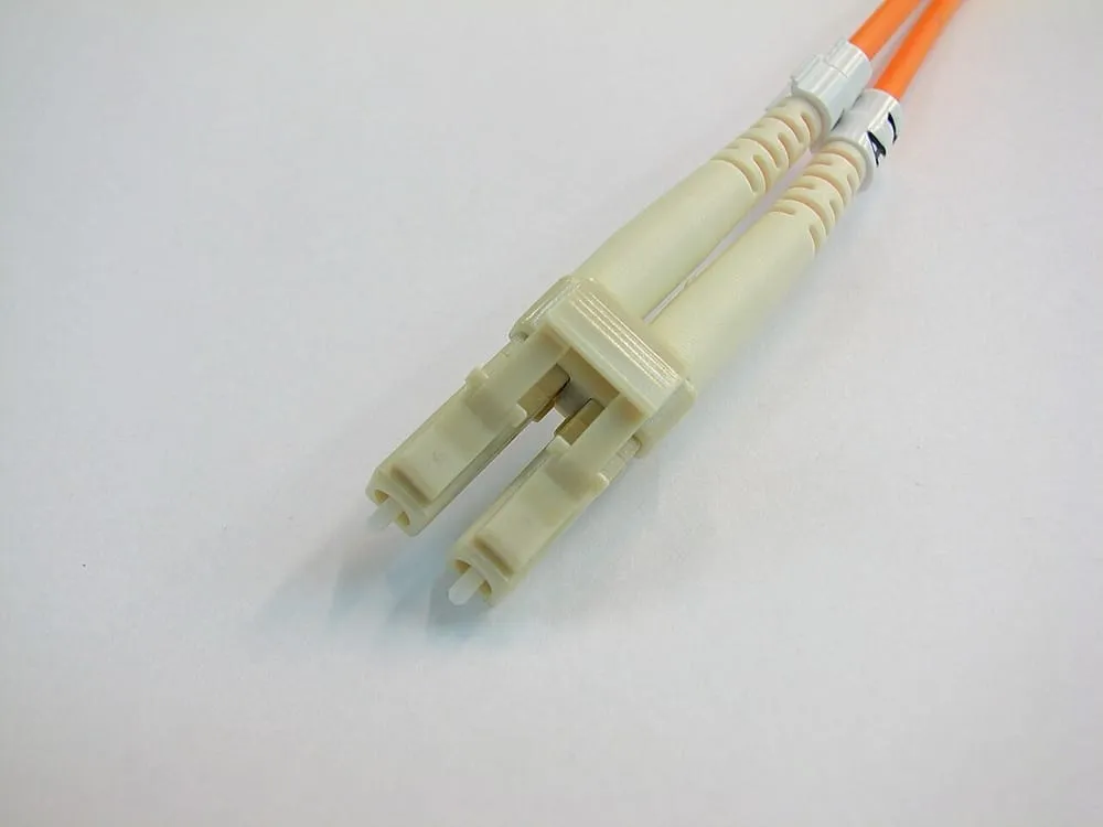A duplex LC connector on an orange multimode patch cable. Two small beige bodies are clipped side by side, each with a slim latch on top like the clip on a telephone plug, and a thin white ferrule protruding from the front of each.