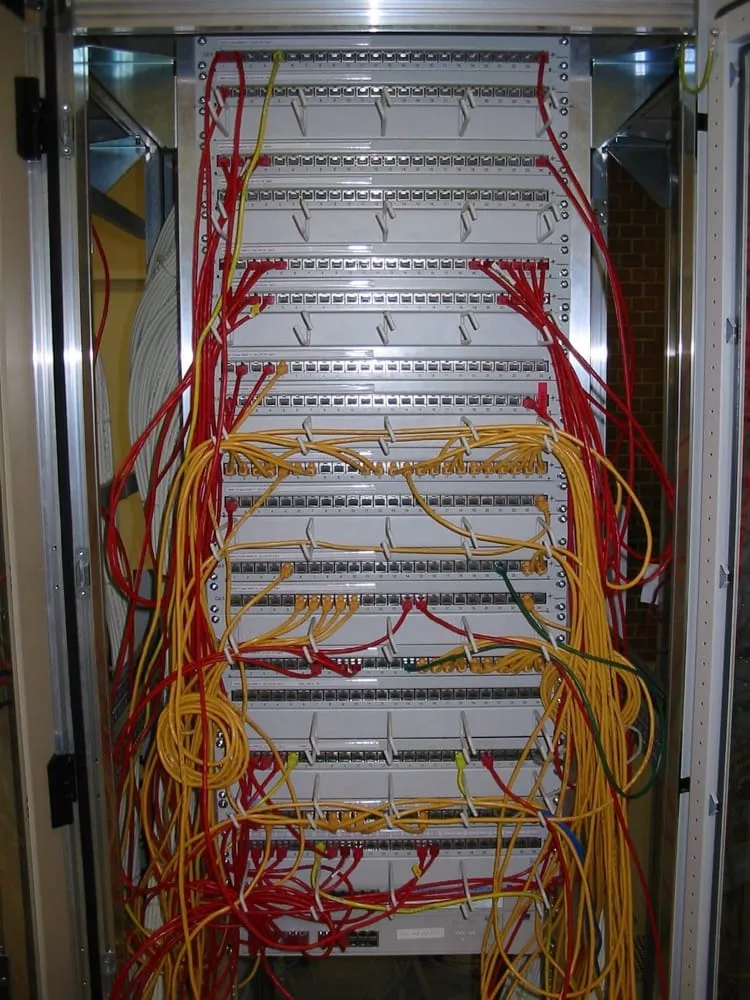 The front of a full rack of patch panels. Row after row of ports fills the frame, with red and yellow patch leads plugged in at scattered positions, looping down the front of the panels and across each other. The leads are not dressed and several hang in long unsupported curves.