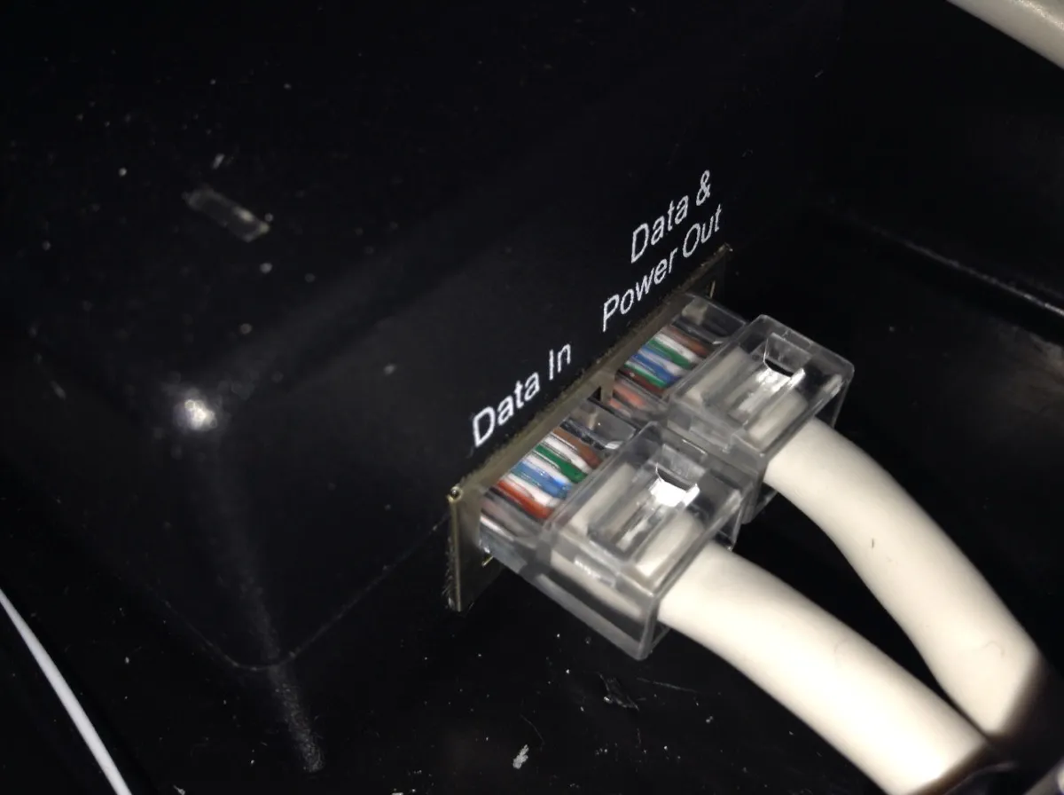 A small black power over Ethernet injector photographed close up. Two sockets sit side by side with transparent-bodied plugs inserted, the individual coloured conductors visible through the housings. The socket on the left is labelled Data In and the one on the right is labelled Data and Power Out.