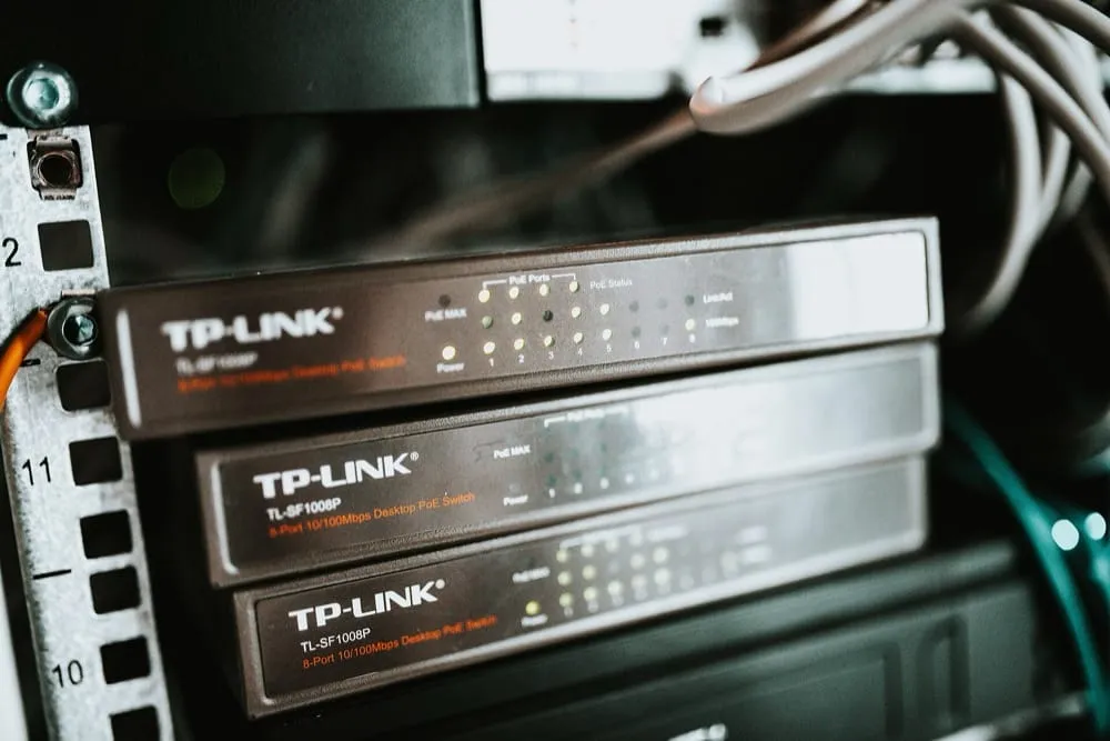 Three identical eight-port TP-LINK PoE switches stacked in a rack, seen from the front. Each has a row of small status lights above the ports, numbered one to eight, with a few lit. The galvanised rack rail on the left is stamped with unit numbers.