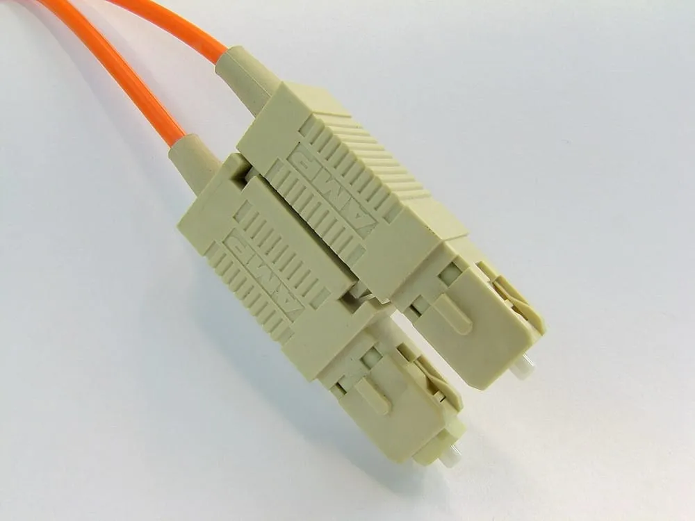 A duplex SC connector on an orange multimode patch cable. Two square beige bodies are clipped side by side, each considerably larger than an LC body, with a ridged grip and a square shroud around a thicker white ferrule.