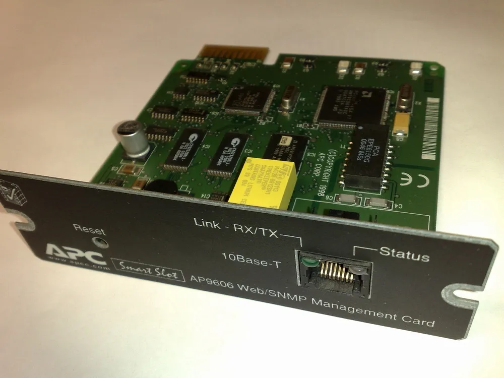 An APC AP9606 Web SNMP Management Card removed from its slot, photographed at an angle. The green circuit board carries several chips, a lithium battery and a barcode. The black front panel is printed with a reset pinhole, a single 10Base-T Ethernet socket with link and status indicators either side of it, and the words AP9606 Web/SNMP Management Card. The board is stamped with a 1998 copyright.
