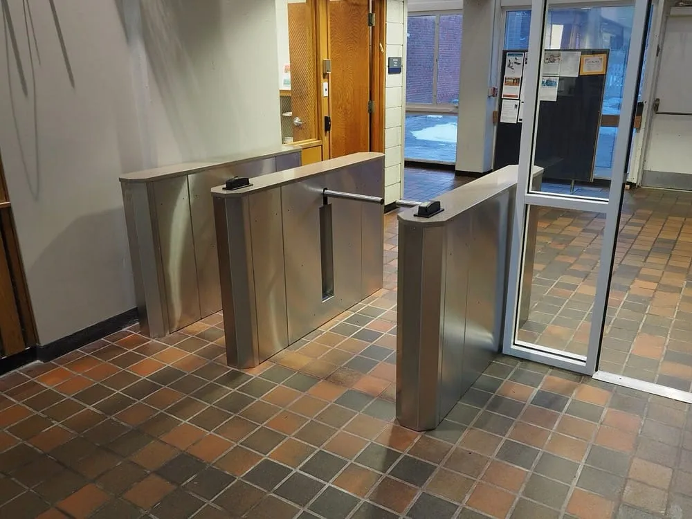 A row of stainless steel speed gates inside a building lobby, each with a small dark card reader mounted on top and a retractable barrier arm, with a glazed entrance door beyond them.