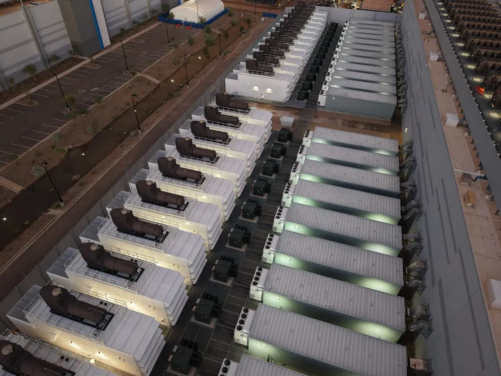 An aerial view at dusk of two long rows of standby generators outside a data centre. Each generator sits in its own white weatherproof enclosure with an exhaust stack and a railed walkway on the roof, and the units are lined up end to end down both sides of a service road. Between and behind them stand fuel tanks and electrical switchgear cabinets, and the windowless wall of the data hall runs down the right of the frame.