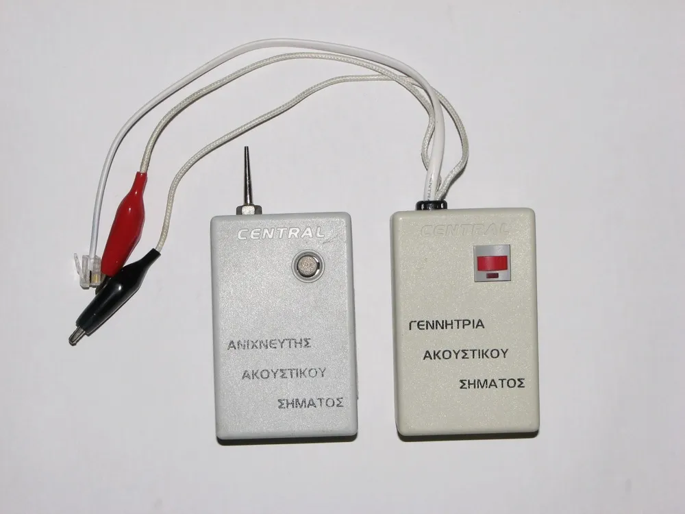 Two small plastic boxes on a white background, each with a white lead. The box on the right is the generator, with a red slide switch, a red and a black crocodile clip and a plug for a network jack on its lead, and a Greek label reading acoustic signal generator. The box on the left is the probe, with a long thin metal spike projecting from the top and a round perforated speaker grille below it, labelled acoustic signal detector.